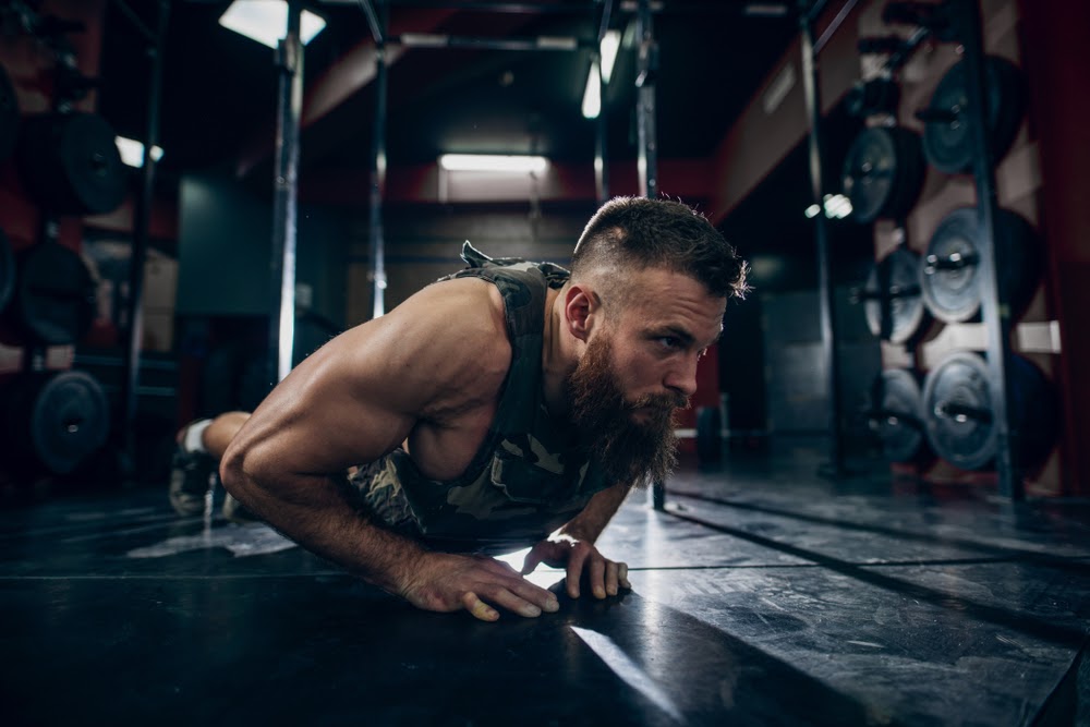 Muscular caucasian bearded man doing push-ups in military style weighted vest in gym. Weight plates in background.